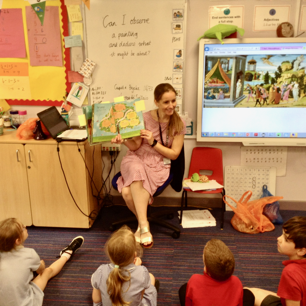 Children sit at the front of a class and look at a class teacher at Hauxton Primary who holds up a picture book about trees next to the Whiteboard with the painting of cupid and psyche from the Fitzwilliam Museum