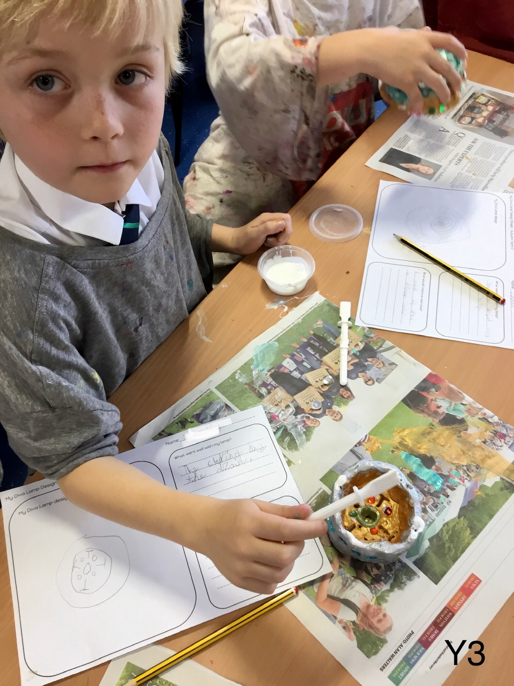 An unknown boy working with glue and paint to decorate a clay pot that he has made with sketchbook pages with his design ideas