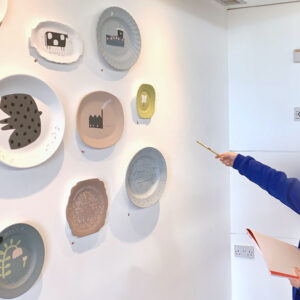 Student points at illustrated plates on the wall in an exhibition by Natalie Deane