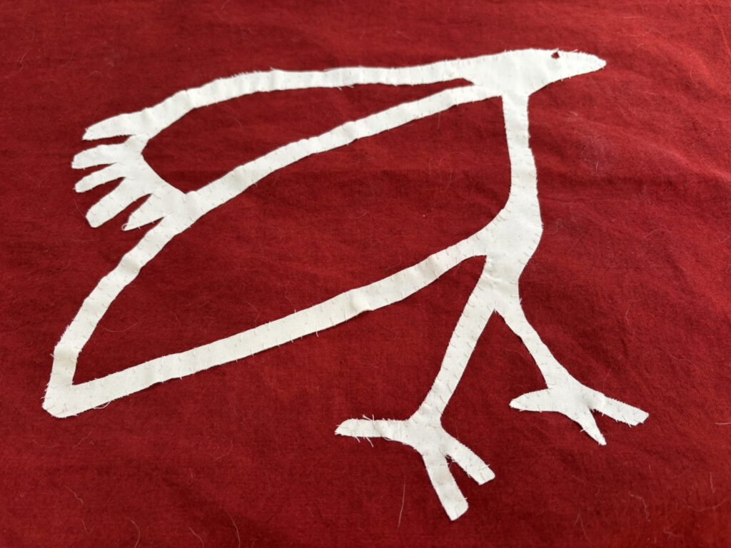 The AccessArt Bird Logo, sewn in white fabric on red cotton
